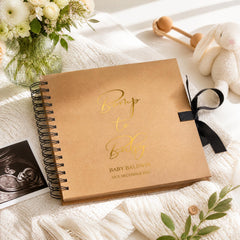 a brown baby announcement book with a gold embossed text, placed on a white surface next to a black and white photograph, a stuffed animal, and a vase of white flowers.