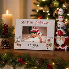 A white picture frame with a photo of a baby and the text "My First Christmas" is displayed on a wooden surface, surrounded by Christmas decorations such as pine cones, a candle, and a small Santa Claus figurine.