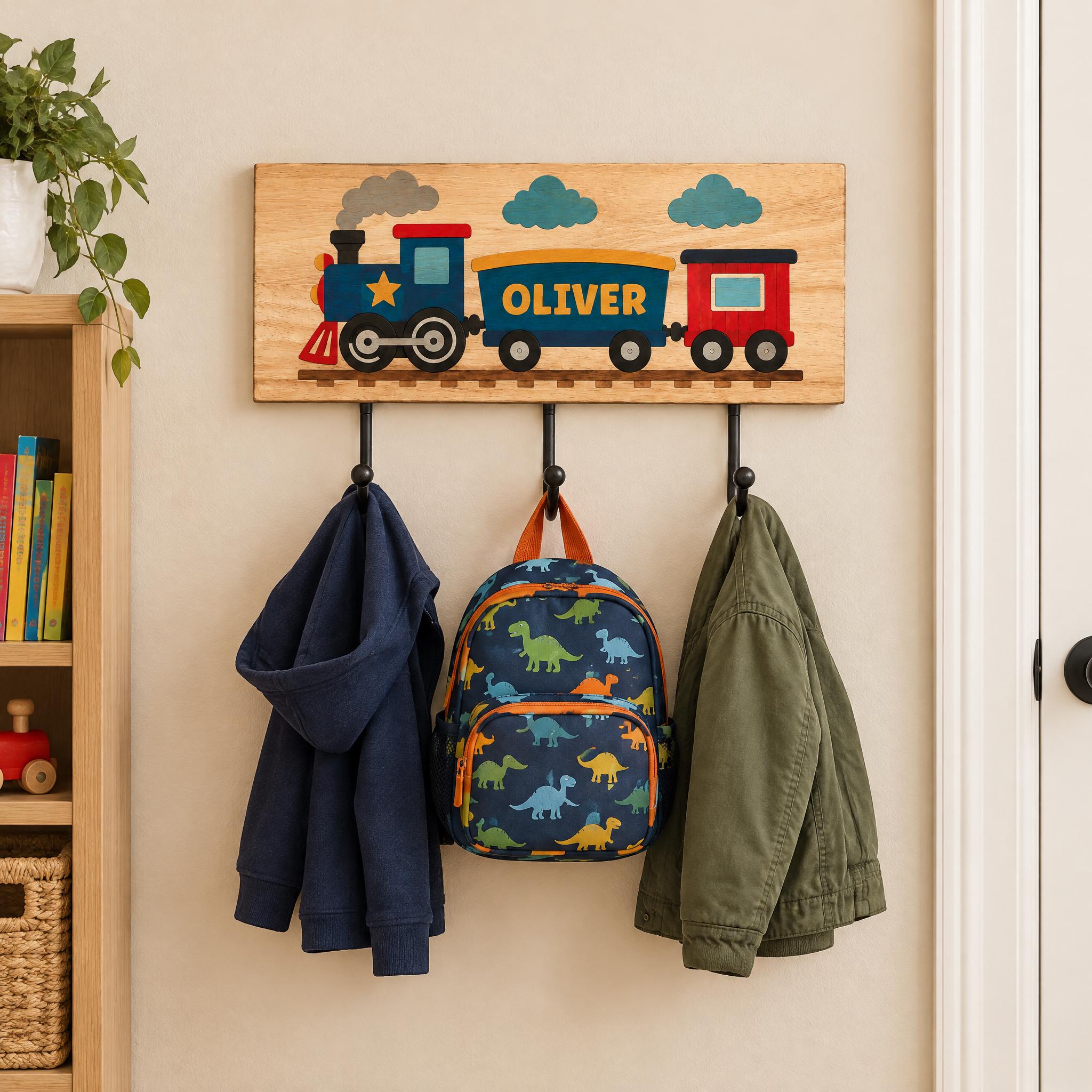 A wooden coat rack with three hooks, displaying a train-themed nameplate, a blue hoodie, and a green jacket.