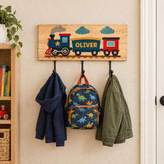 A wooden coat rack with three hooks, displaying a train-themed nameplate, a blue hoodie, and a green jacket.
