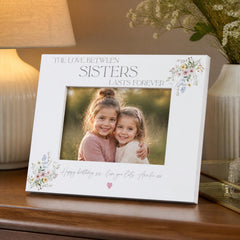 A white picture frame with a photo of two young girls, likely sisters, smiling and embracing each other. The frame has a floral design and the text "The love between sisters lasts forever" written on it.
