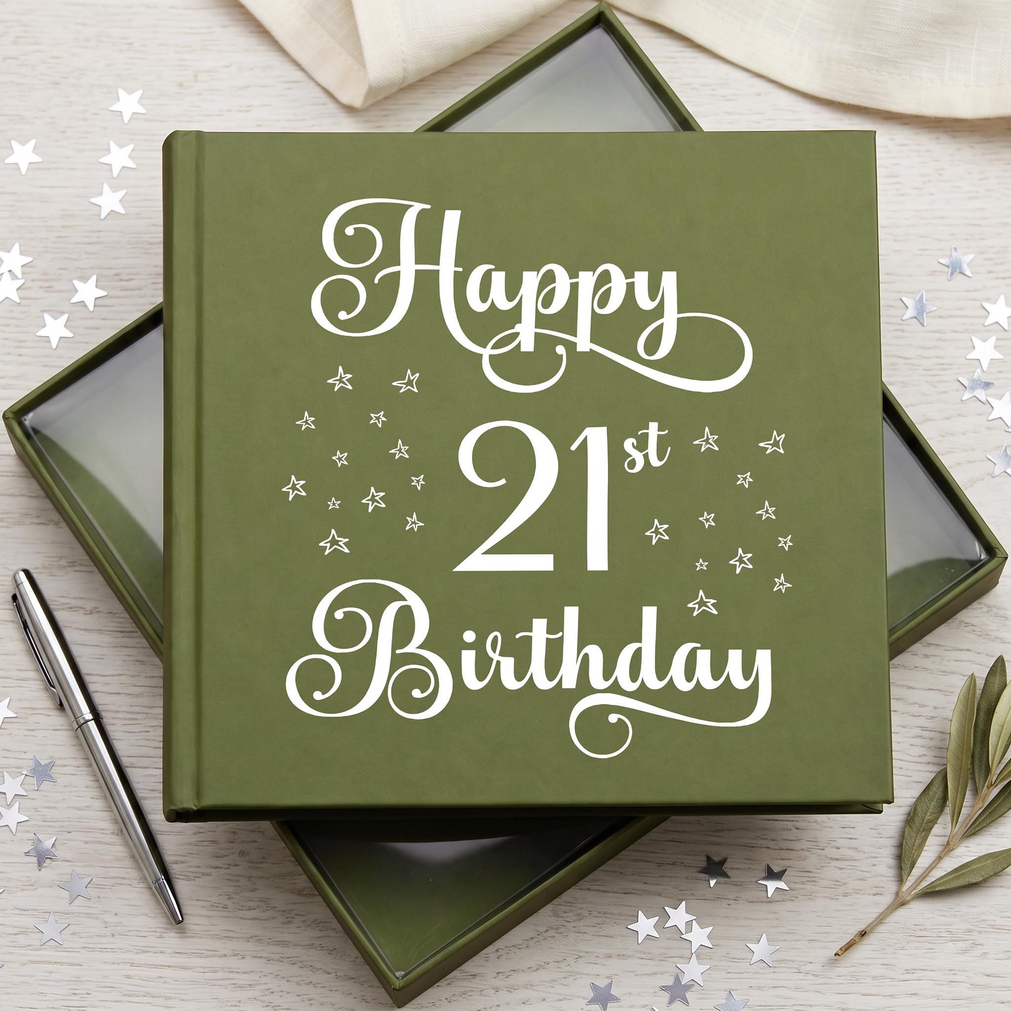 A green book titled "Happy 21st Birthday" with white stars on the cover, placed on a wooden surface surrounded by scattered silver stars.