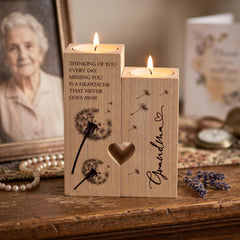 a wooden candle holder with a heart-shaped cutout, a dandelion design, and a message that reads "Thinking of you every day, missing you is a heartache that never goes away". The candle holder is placed on a wooden surface, with a framed photo of an elderly woman in the background.