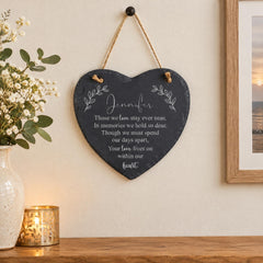 a heart-shaped sign hanging on a wall, with a vase of white flowers and a candle nearby.