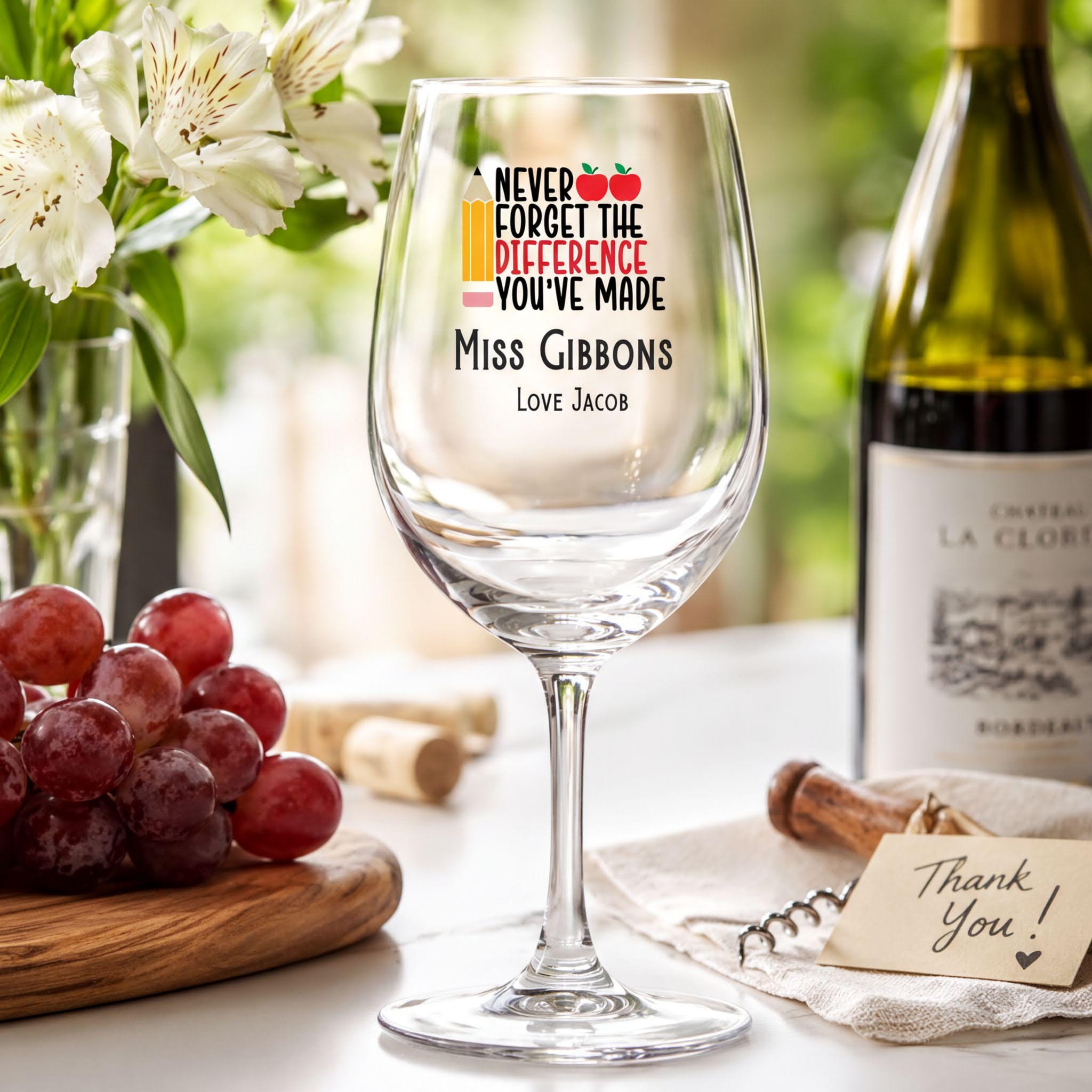 a wine glass with a personalized message, a bottle of wine, and a wooden cutting board with grapes and a corkscrew.