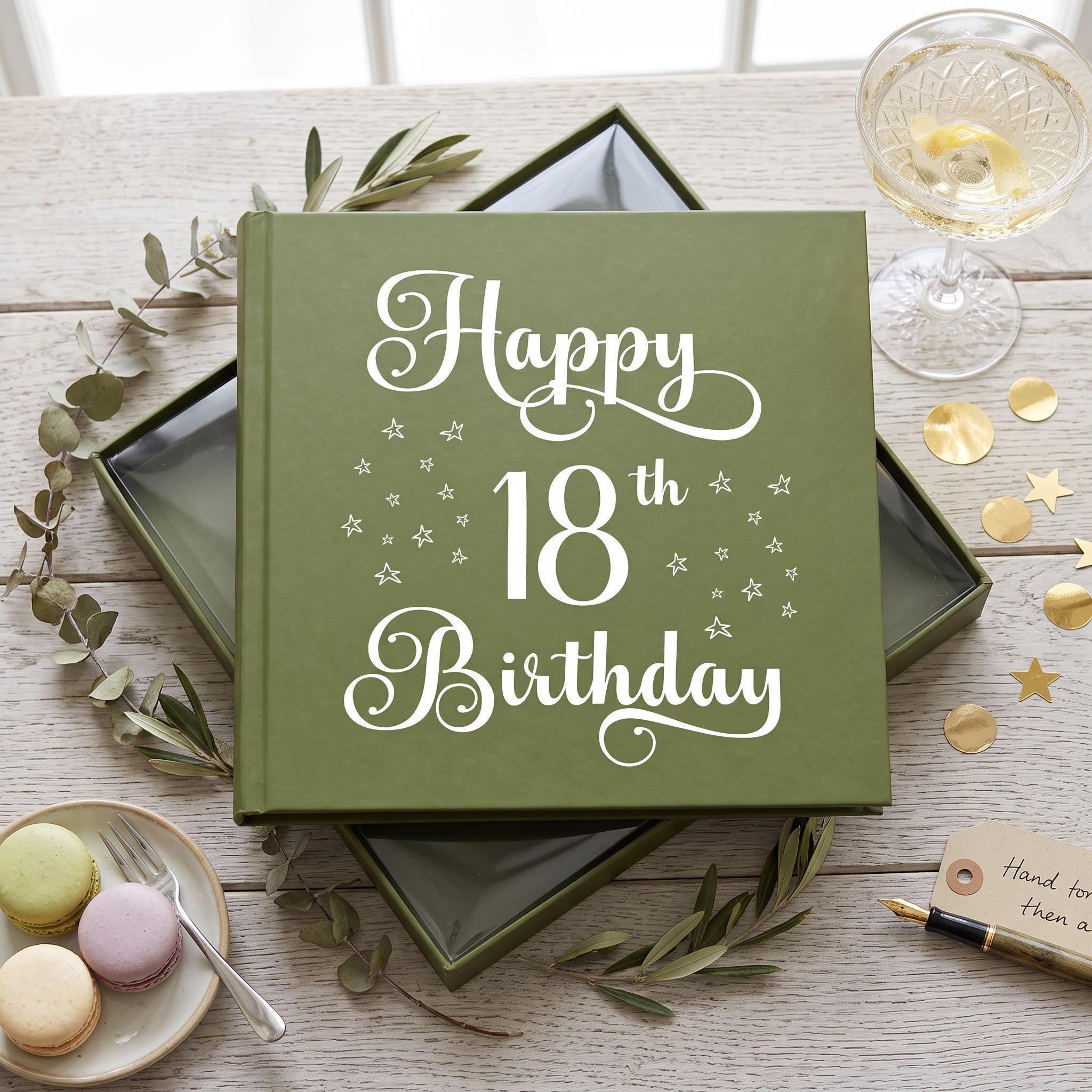 A green book titled "Happy 18th Birthday" sits on a wooden table, surrounded by a glass of wine, a plate of macarons, and some gold stars.