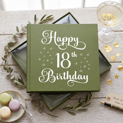 A green book titled "Happy 18th Birthday" sits on a wooden table, surrounded by a glass of wine, a plate of macarons, and some gold stars.