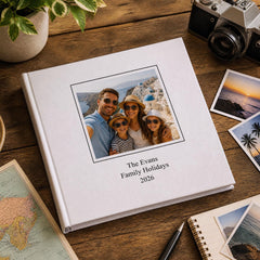 a white photo album with a family photo on the cover, placed on a wooden surface alongside a camera, a map, and some photographs.