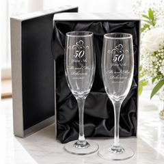 a pair of clear champagne glasses with a black box and a vase of white flowers in the background.