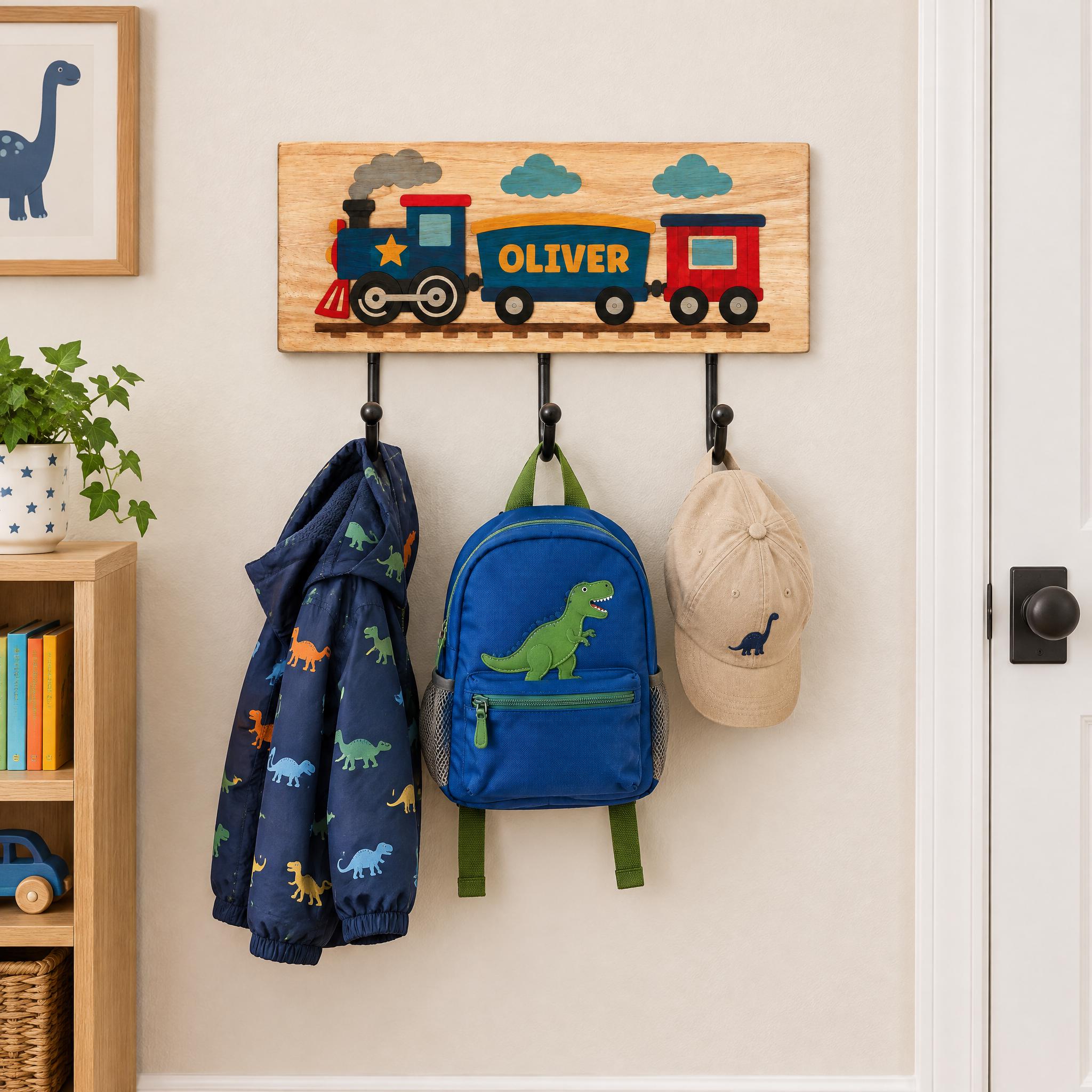 A wooden coat rack with a train-themed sign hanging on it, displaying the name "OLIVER" and a train engine. The coat rack also holds a blue backpack with a dinosaur design and a beige baseball cap.