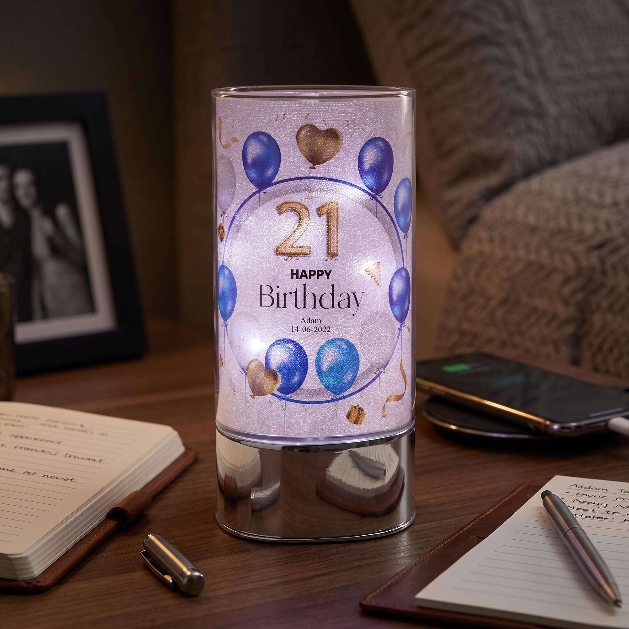 A birthday-themed lighted candle holder sits on a wooden table, surrounded by various items such as a notebook, pen, and smartphone.
