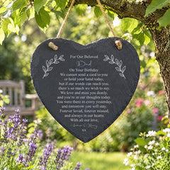 A heart-shaped slate with a heartfelt message for a loved one, hanging on a tree branch in a garden setting with purple flowers in the background.
