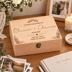 A wooden box with a personalized wedding memory keepsake, featuring the names "Oliver & Claire" and the date "30.06.2026" along with the phrase "The day our forever began". The box is placed on a wooden surface, surrounded by wedding-related items such as a candle, photographs, and a notebook.