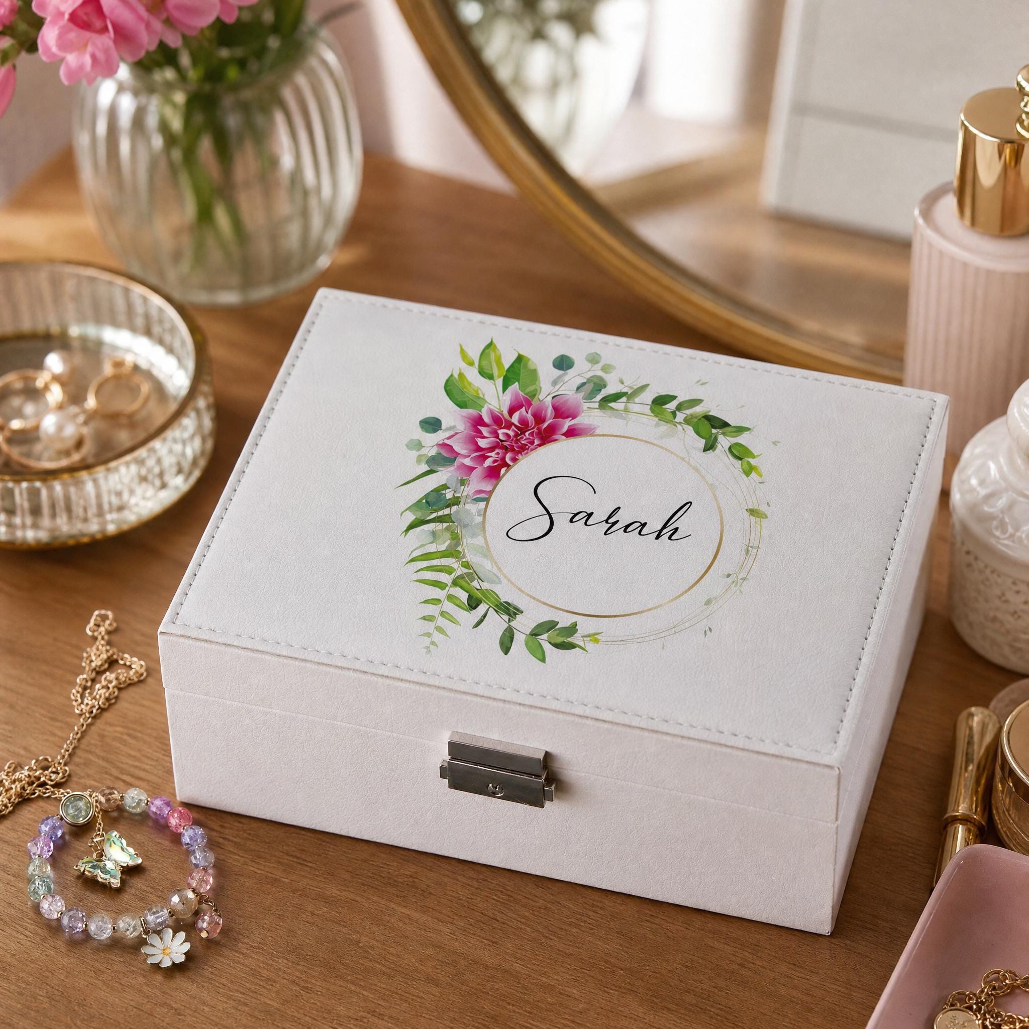 A white jewelry box with a floral design and the name "Sarah" printed on it, placed on a wooden surface alongside other personal items such as a bracelet and a vase with flowers.