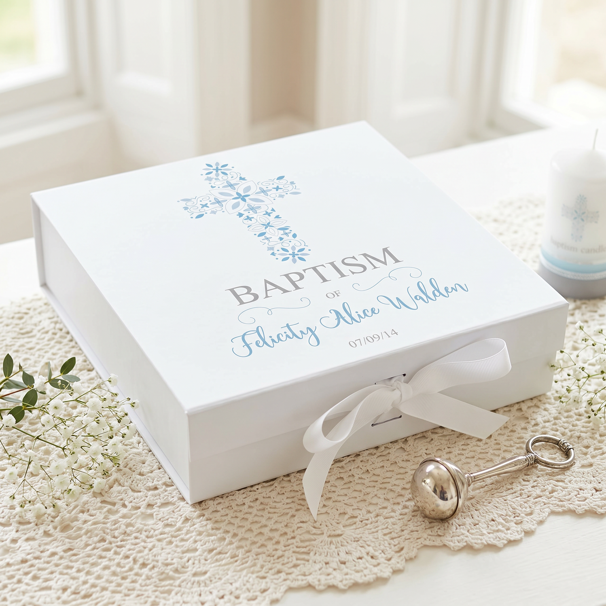 A white gift box with a blue cross and floral design, placed on a lace doily. Next to the gift box is a silver baby rattle.