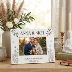 A white picture frame with a photo of a couple, surrounded by decorative elements such as a vase of flowers, candles, and a book.