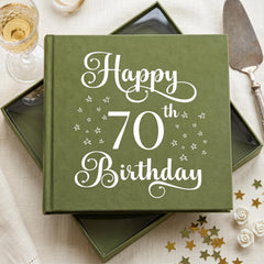 a green book with the text "Happy 70th Birthday" printed on it, along with white stars. The book is placed on a table with a wine glass, a cake, and some gold stars scattered around it.