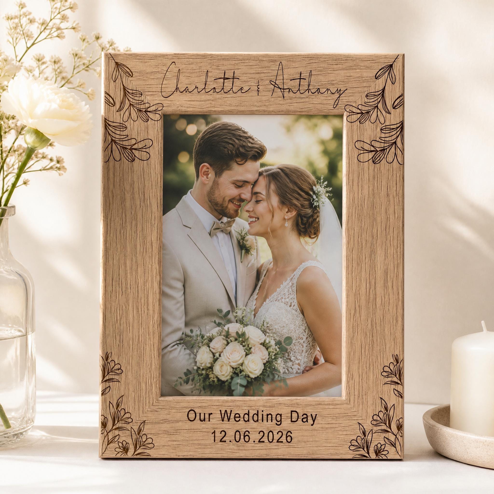 A wooden picture frame with a photo of a couple, presumably a bride and groom, inside it. The frame has a floral design around the edges and the names 'Charlotte & Anthony' written on it. The photo appears to be a wedding picture, and the date '12.06.2026' is written below the frame. The frame is placed on a table, with a vase of flowers and a candle nearby.