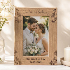 A wooden picture frame with a photo of a couple, presumably a bride and groom, inside it. The frame has a floral design around the edges and the names 'Charlotte & Anthony' written on it. The photo appears to be a wedding picture, and the date '12.06.2026' is written below the frame. The frame is placed on a table, with a vase of flowers and a candle nearby.