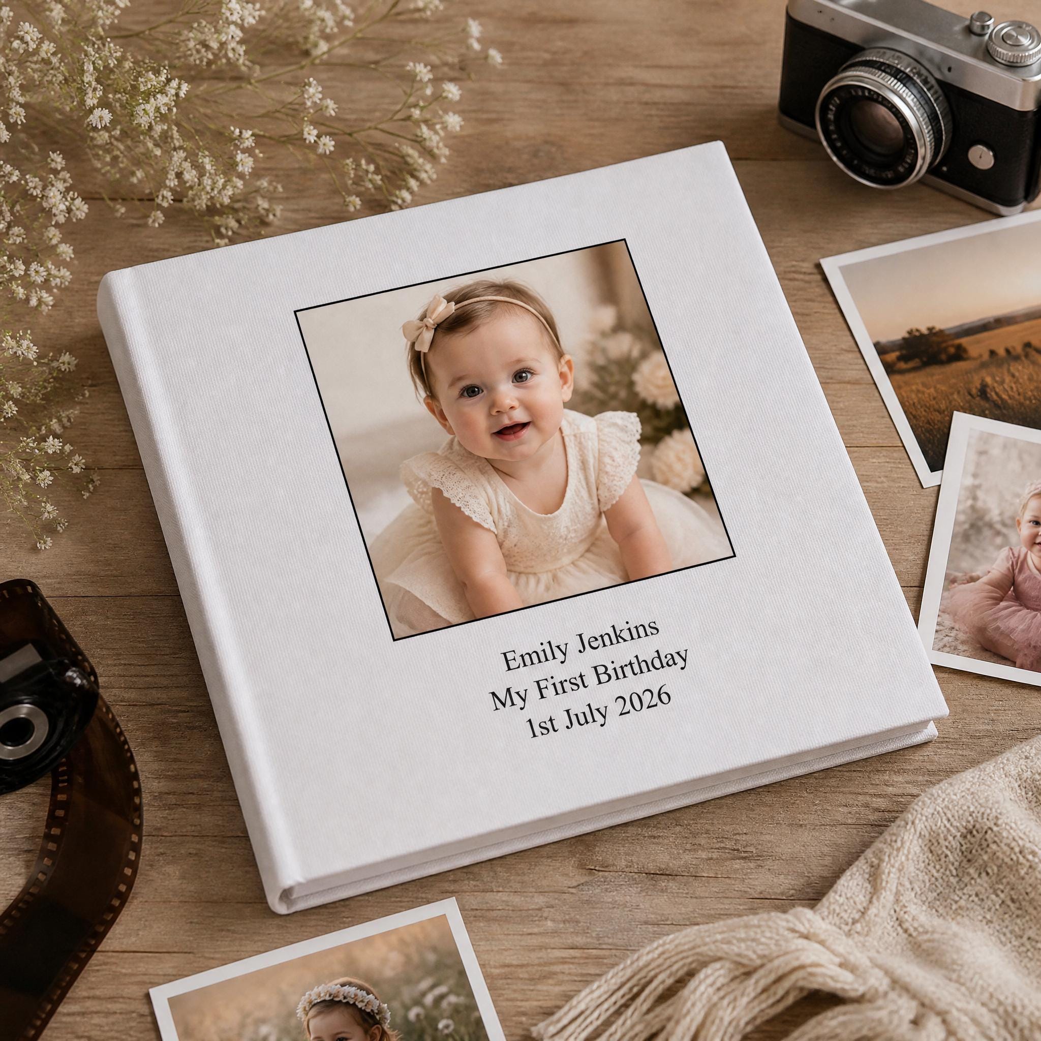 A white photo album with a picture of a smiling baby girl on the cover, placed on a wooden surface alongside other items such as a camera and some flowers.