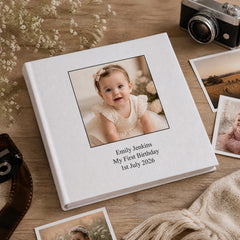 A white photo album with a picture of a smiling baby girl on the cover, placed on a wooden surface alongside other items such as a camera and some flowers.