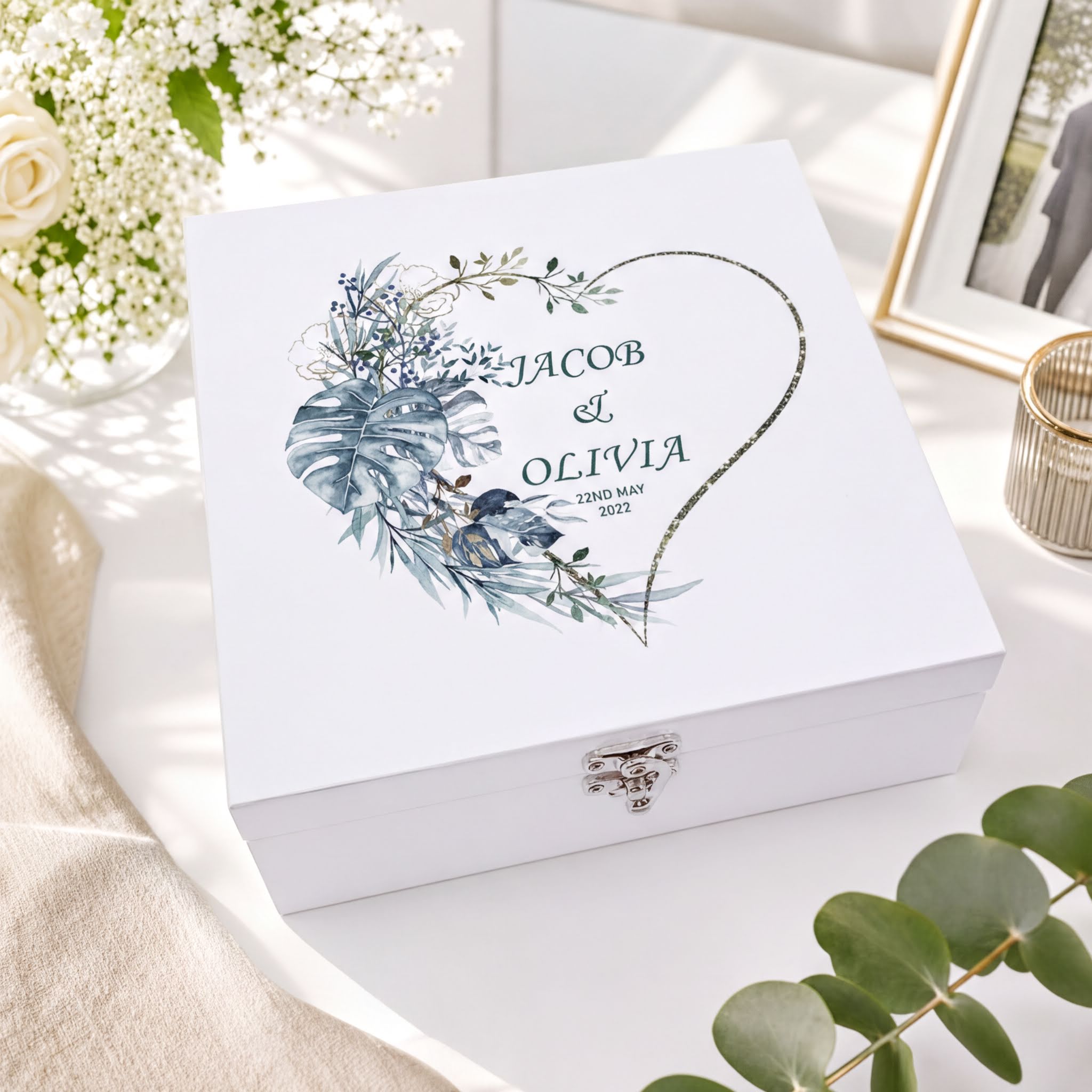 A white box with a floral design and the names " Jacob & Olivia" printed on it, placed on a table with a vase of white flowers and a framed photo.