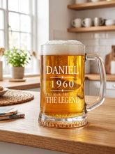 A glass mug filled with beer sits on a wooden table, with a potted plant and kitchen items visible in the background.