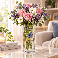 A glass vase filled with pink roses, white daisies, and purple flowers sits on a marble table, with a candle and a stack of books nearby.