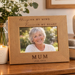 A wooden picture frame with a photo of an older woman inside, placed on a wooden surface. The frame has a message engraved on it that says "Always on my mind Forever in my heart" and "MUM 1946-2026".