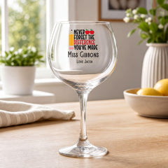 A wine glass with a personalized message on it, placed on a wooden table in a room with a potted plant and a bowl of fruit in the background.