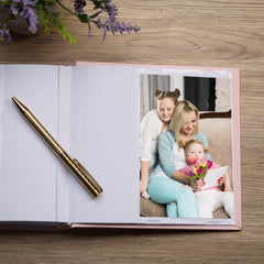 an open photo album with a photograph of three people, including a woman and two children, sitting together on a couch. A pen is placed next to the open album, and there are purple flowers on the wooden surface.