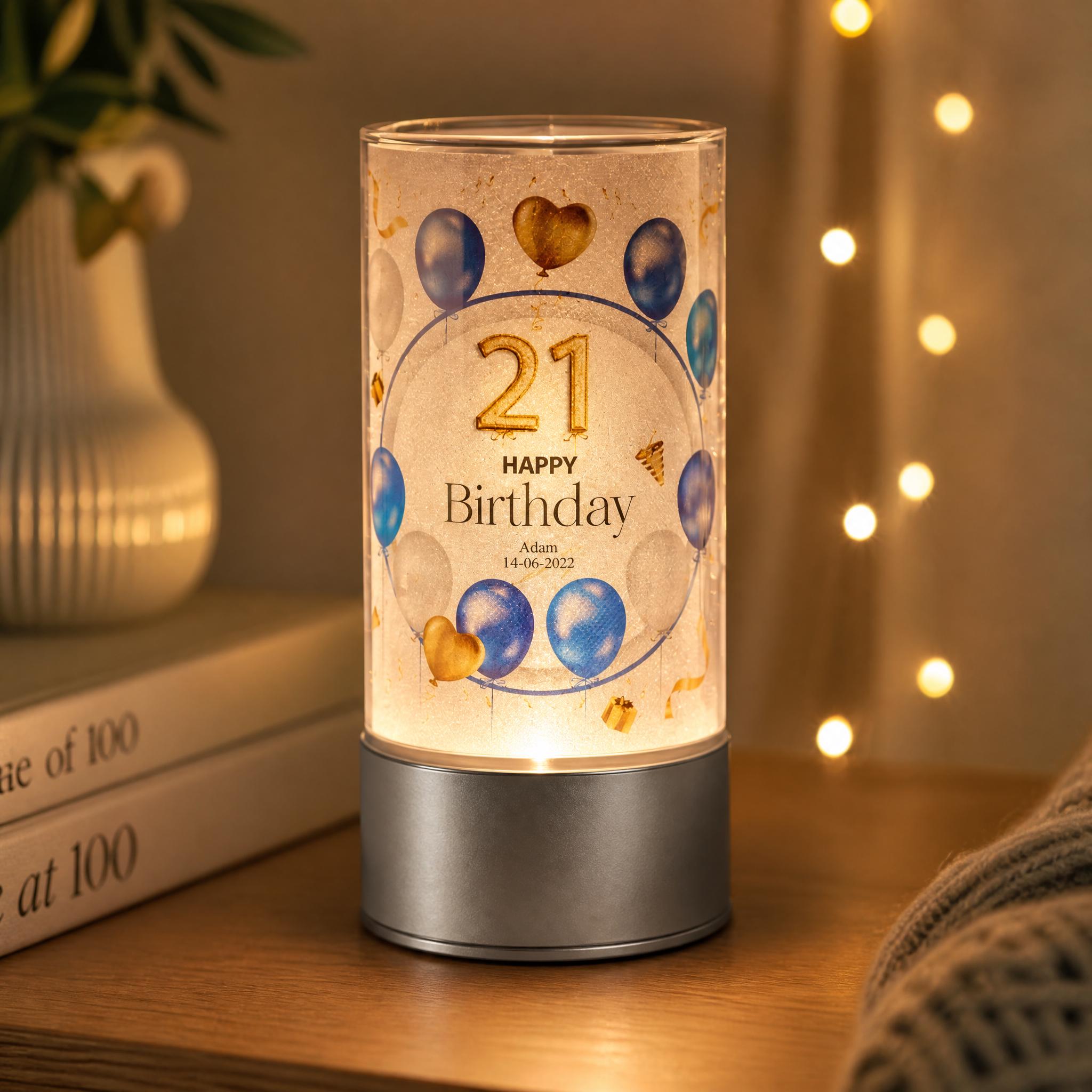 A birthday-themed lighted lamp with the number 21 and balloons on its surface, placed on a wooden surface next to a stack of books and a plant.