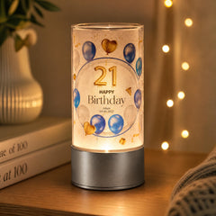 A birthday-themed lighted lamp with the number 21 and balloons on its surface, placed on a wooden surface next to a stack of books and a plant.