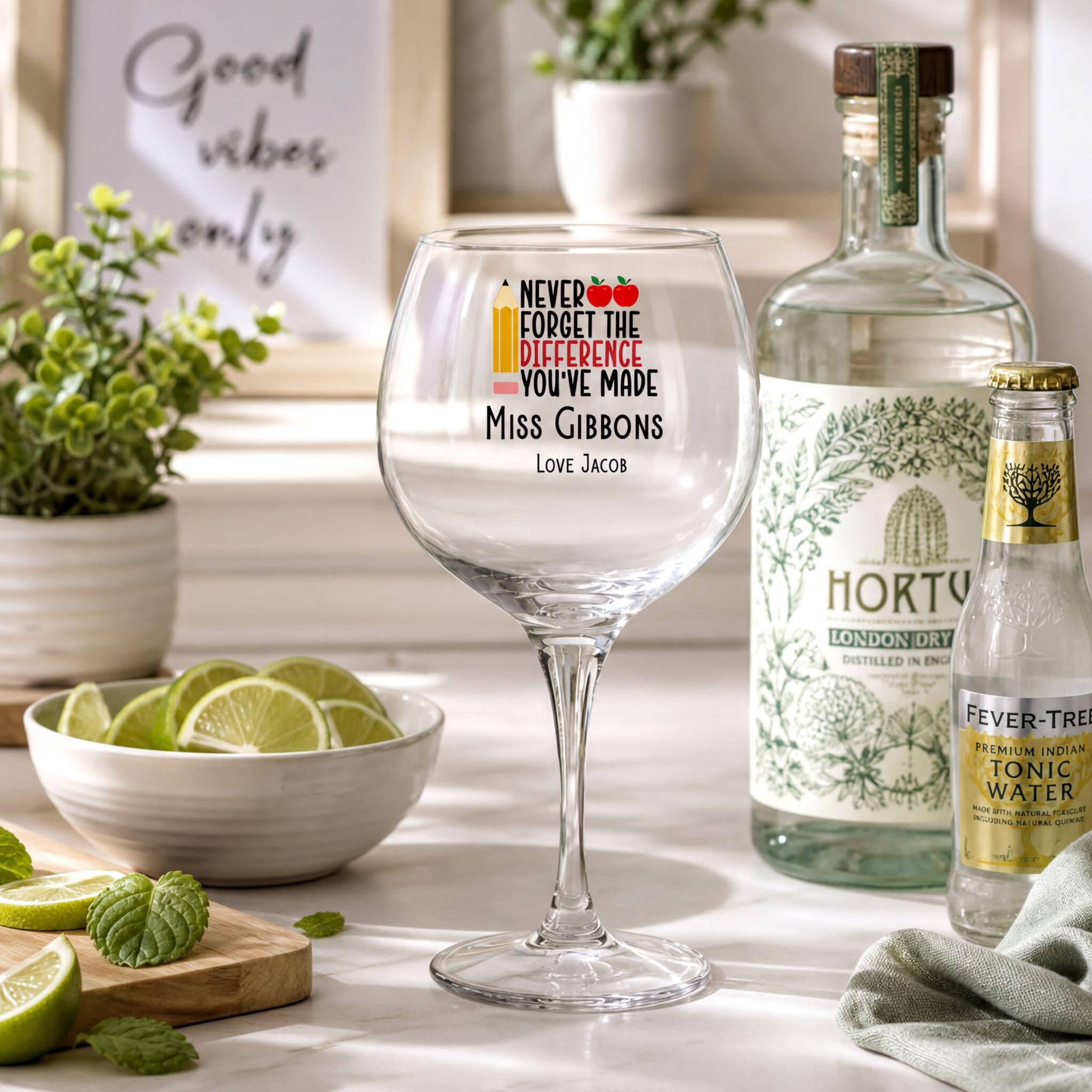 a table with a wine glass, a bottle of gin, a bowl of limes, and some plants. The wine glass has a sticker on it with a message about gin.