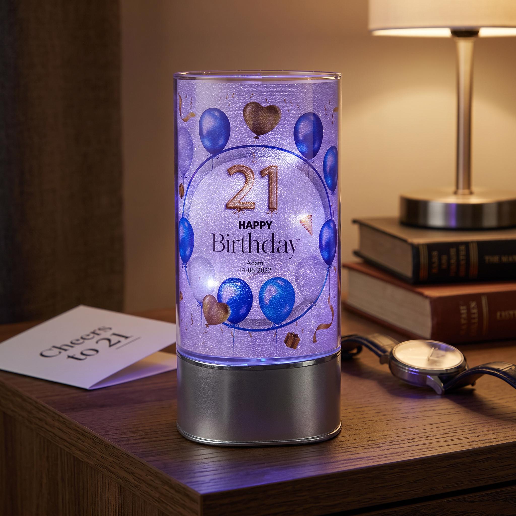 A birthday-themed lighted candle holder with a purple background and blue balloons, placed on a wooden surface next to a lamp and some books.