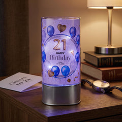 A birthday-themed lighted candle holder with a purple background and blue balloons, placed on a wooden surface next to a lamp and some books.