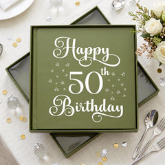 A green box with a white and gold design, displaying the text "Happy 50th Birthday" surrounded by stars. The box is placed on a table with a white tablecloth, and there are silverware and a bouquet of flowers nearby.