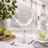 a wine glass with a personalized message, a bottle of gin, and some fruits and a spoon on a table.