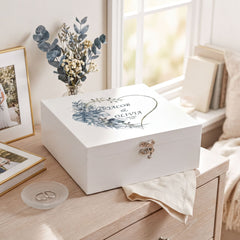 A white wooden box with a floral design sits on a wooden dresser, accompanied by a vase of flowers, a framed photo, and a book.