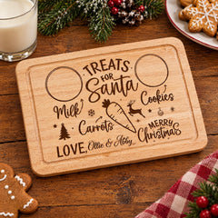 A wooden cutting board with a personalized message engraved on it, surrounded by holiday decorations and treats, including cookies and a gingerbread man.