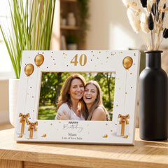 A white picture frame with gold balloons and bows, displaying a photograph of two women smiling. The frame has the number "40" and the text "Happy Birthday" written on it. The frame is placed on a wooden surface, with a vase of flowers and a potted plant in the background.