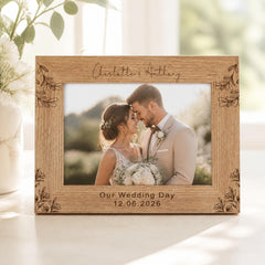 A wooden picture frame with a photo of a couple embracing, surrounded by floral designs. The frame has a personalized inscription that reads "Our Wedding Day 12.06.2026".