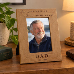 A wooden picture frame with a photo of an older man smiling, placed on a wooden surface next to a book and a plant.