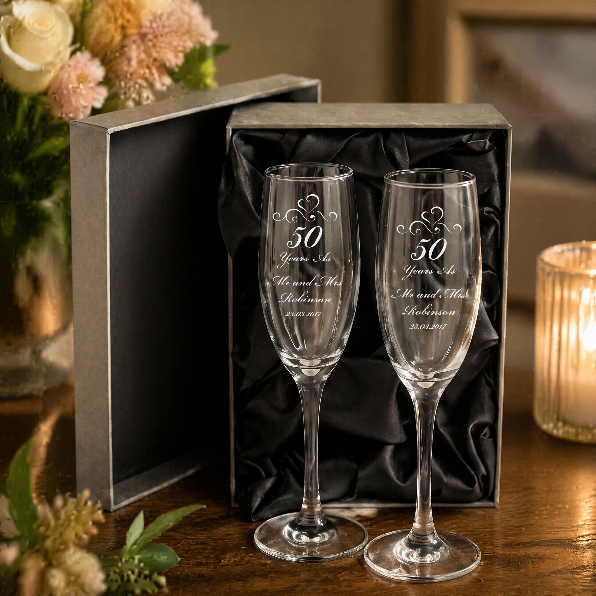a pair of elegant champagne glasses, each inscribed with a 50th anniversary message, placed on a wooden table. The glasses are accompanied by a black box, and a vase of flowers is visible in the background.