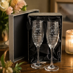 a pair of elegant champagne glasses, each inscribed with a 50th anniversary message, placed on a wooden table. The glasses are accompanied by a black box, and a vase of flowers is visible in the background.
