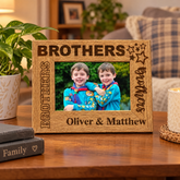 A wooden picture frame with the text "BROTHERS" and "Oliver & Matthew" engraved on it, displaying a photo of two smiling boys. The frame is placed on a wooden table, with a potted plant, a book, and a candle nearby.