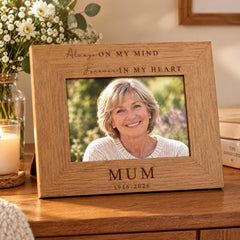 A wooden picture frame with a photograph of a smiling woman inside, placed on a wooden surface. The frame has a message engraved on it that reads "Always on my mind Forever in my heart".