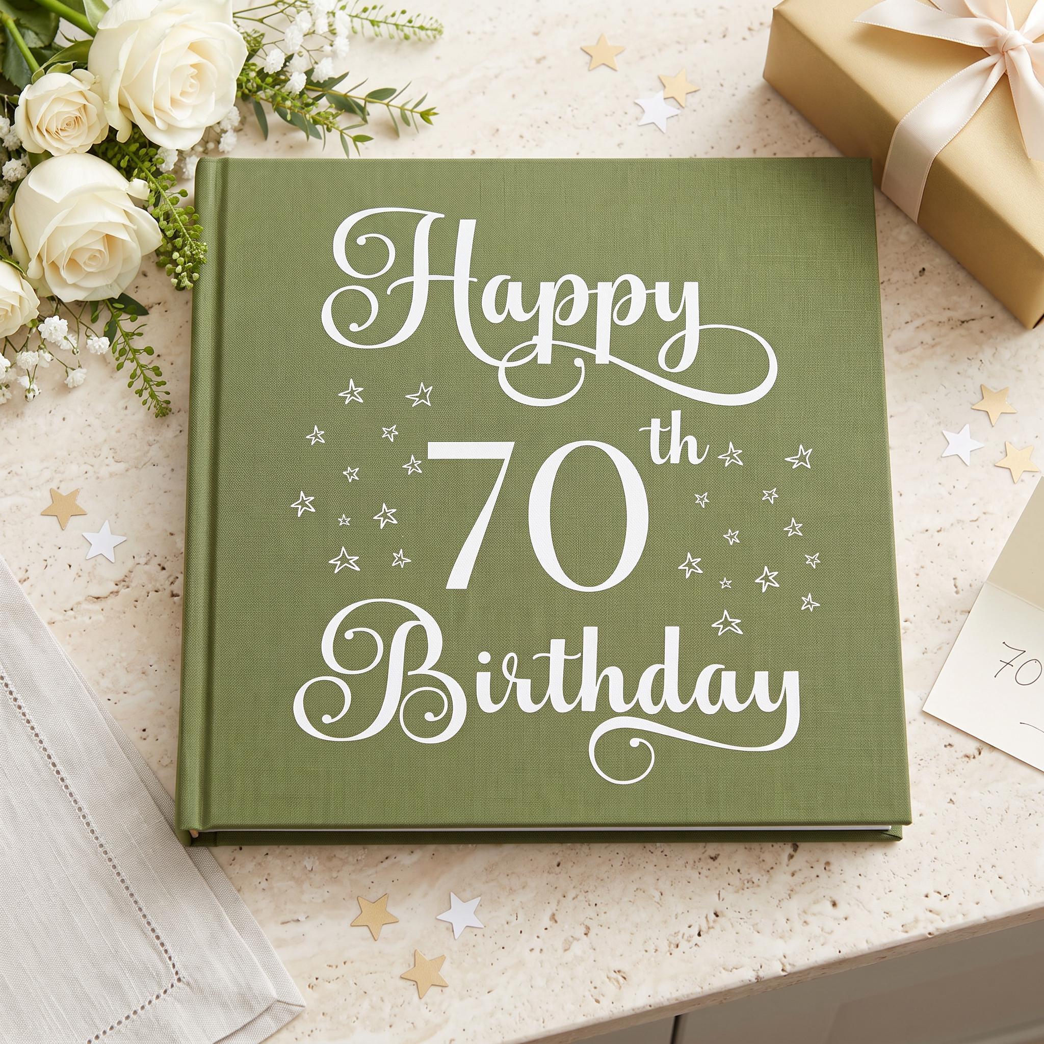 A green book titled "Happy 70th Birthday" with white text and stars on the cover, placed on a table with white flowers and a gift box.