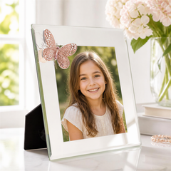 A young girl with long brown hair is smiling at the camera, framed in a white picture frame with a pink butterfly decoration. The frame is placed on a table, with a vase of white flowers and a stack of books nearby.