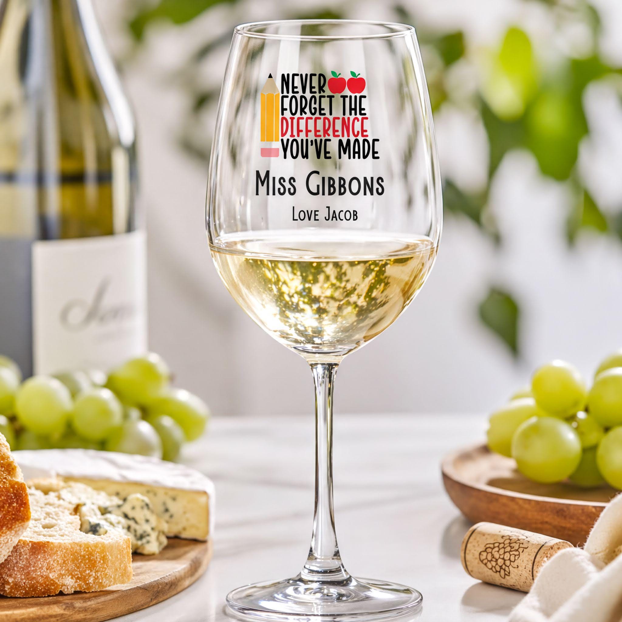 a wine glass with a personalized message, a bottle of wine, and some cheese and grapes on a table.